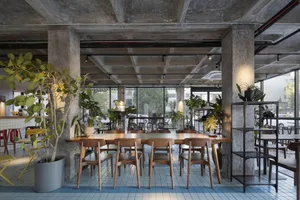 Modern restaurant interior with a large wooden dining table surrounded by metal chairs, potted plants, and vases, under natural light