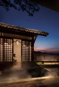 A woman sits in front of a doorway in a traditional Asian-style house with a wooden exterior, surrounded by a calm and serene atmosphere