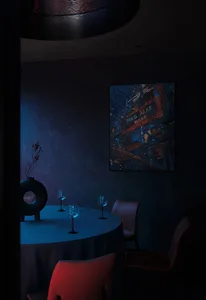 A dark and intimate dining area with a round table, chairs, and wine glasses, set for dinner service