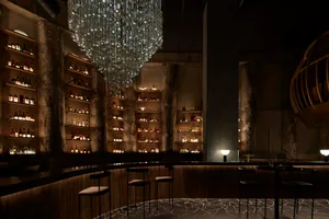 A sophisticated bar area with a wooden counter, stools, and a large chandelier, surrounded by bottles and a modern ambiance
