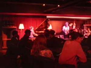 A warm and intimate indoor event space with a stage and audience seated in chairs, surrounded by red walls and a piano, with musicians playing instruments