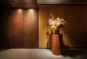 A cozy hallway with a yellow wall, a wooden vase with flowers, and a wooden stand, with an open door revealing a room beyond