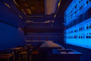 A dimly lit bar area with a sleek counter, white and blue elements, and chairs for seating, creating a cozy atmosphere for guests