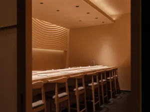 A casual restaurant dining area with wooden tables and chairs, set against a warm tan-colored wall backdrop