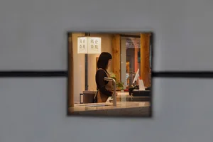 A waitress stands behind a counter in a casual dining restaurant with potted plants and two cups on the counter