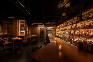 A warm and inviting bar area with a long wooden counter, multiple chairs, and bottles displayed, set in a restaurant
