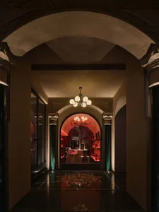 Ornate hallway with red walls, a grand archway, and chandeliers providing illumination