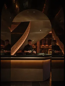 A lively bar scene with two bartenders serving customers, surrounded by bottles, cups, and wine glasses on a wooden counter in a restaurant