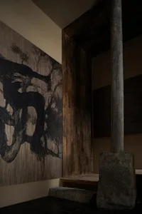 A darkened room features an abstract painting of a tree on the wall, created with wood and paint, in predominantly black and brown hues, with a moody atmosphere