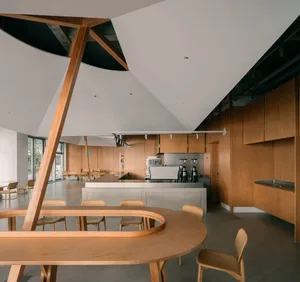 Open modern kitchen and dining area with wooden cabinets, countertops, and a large circular dining table, featuring potted plants and a refrigerator, oven, and two sinks