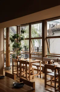 Modern restaurant interior with wooden tables and chairs, large windows, and potted plants, featuring a welcoming atmosphere