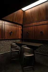 A modern restaurant or bar area with wooden walls and floors, featuring two black leather chairs and a marble dining table, well-lit with natural or artificial light