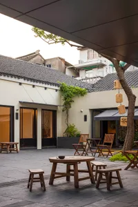 A welcoming courtyard dining area with wooden chairs, a dining table, and potted plants, set within a larger building complex