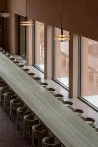 A well-lit restaurant with a long dining table, chairs, and natural light from windows