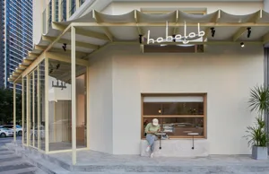 白色店面配黃色遮陽篷與盆栽，女子坐在外頭長椅上，背景有停放的汽車