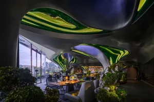 A modern dining area with a large table and chairs, surrounded by potted plants and a kitchen in the background