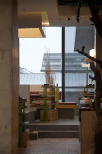 A well-lit restaurant dining area with tables, chairs, potted plants, and vases, surrounded by windows allowing natural light to enter