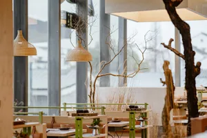 A modern restaurant dining area with tables, chairs, and potted plants, set for guests with various tableware and utensils