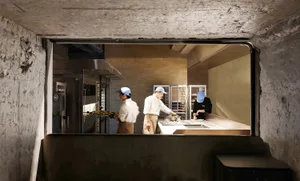 Three men in blue hats work together in a kitchen with an oven, sink, and refrigerator, surrounded by bowls and focused on meal preparation
