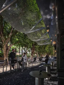 A casual outdoor seating area with metal chairs and tables under a large umbrella, surrounded by neutral tones and natural light