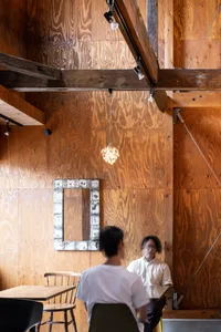 A man sits in front of a mirror on a wooden wall with a rustic feel, surrounded by two people in a cozy living or bedroom space