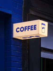 Exterior view of a coffee shop with a brightly lit sign featuring a cup and spoon, surrounded by city buildings
