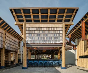 An open-air restaurant with a wooden roof, a bar area, and multiple chairs, surrounded by people enjoying a relaxed atmosphere