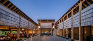 An open-air restaurant with a wooden structure and thatched roof, featuring a dining area with chairs and tables in a tropical setting
