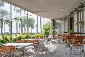 A restaurant dining area with modern and traditional elements, featuring multiple tables, chairs, potted plants, and palm trees in the background, creating a comfortable and inviting atmosphere