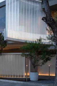 A modern building with a white concrete facade and a large window, surrounded by two trees and natural light