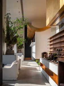 A modern restaurant interior with white and brown tones, featuring a dining area with multiple tables and chairs, a bar area with decorative items, and potted plants