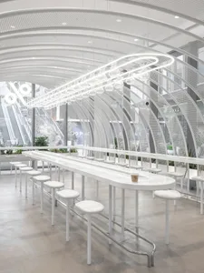 A modern restaurant interior with a long white table surrounded by chairs, potted plants, and cups on the tables
