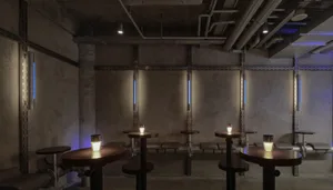 A modern bar area with a long counter, stools, dining tables, and a mix of stone and concrete walls, with various lighting sources creating a stylish ambiance
