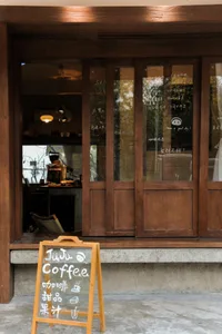 A cozy coffee shop with wooden walls, a window display, and comfortable seating areas, featuring a dining table and couch, with cups and bowls scattered throughout the scene