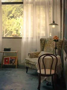 Cozy living space with a chair, cushion, and dining table, surrounded by books and a vase, in front of a window