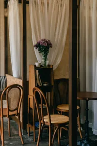Cozy living space with a wooden floor, a dining table, chairs, and a vase with flowers on a wooden stand, set in a warm and inviting atmosphere