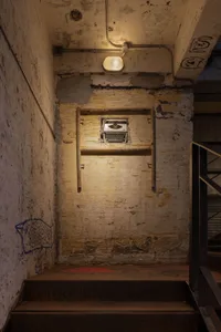 A narrow, dimly-lit hallway with a yellow wall, concrete surfaces, and a small window allowing natural light