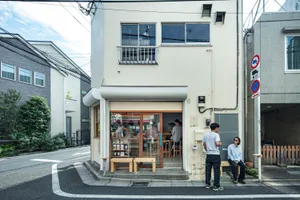 熱鬧的都市街景，一棟高聳的白建築帶有陽台，周圍環繞著人群與車輛，擺放盆栽，充滿生氣氛圍