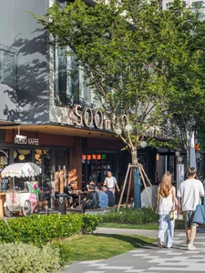 An inviting outdoor cafe area with wooden benches, tables, and umbrellas, surrounded by trees and bustling with people
