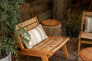 A wooden bench with a woven design, situated in front of a tree trunk, with two pillows and a natural atmosphere
