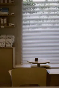 A modern living room with a dining table, couch, chairs, and various items scattered throughout, with a window with white blinds