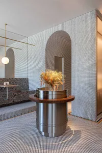 A warm and inviting living area with a wooden table, metal vase with flowers, and a dining table, surrounded by a tile-adorned wall and two chairs