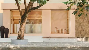 A building with a pink facade features a large window and seating options, including two benches and three chairs, in a calm and inviting atmosphere
