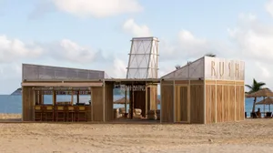 An open-air restaurant on a beach with a wooden structure, chairs, tables, and bottles, surrounded by a relaxed atmosphere