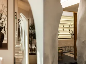 A minimalist living space with white surfaces, potted plants, vases, and chairs, featuring a large window and a hallway leading to a door