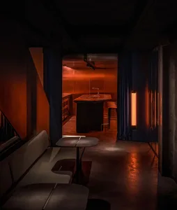 Dimly lit bar or restaurant space with a dining table, chairs, and a kitchenette, featuring wooden walls and a cozy ambiance