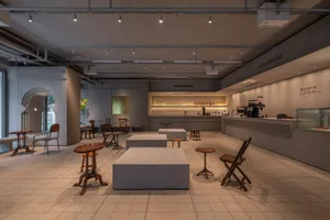 Modern restaurant with exposed cement walls, a central bar, and scattered chairs and dining tables, with a potted plant adding a touch of greenery