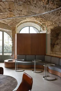 Cozy living area with stone walls, wooden furniture, and a large window, featuring a couch and several chairs around a central dining table