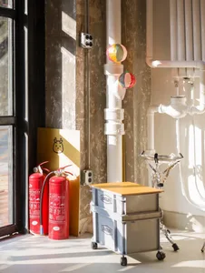 A cluttered living area with a window, red fire extinguishers, bottles, and a bowl, suggesting a functional yet somewhat disorganized space