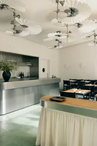 Modern restaurant kitchen with stainless steel appliances, wine glasses, and a doorway to a spacious dining hall with chairs and dining tables